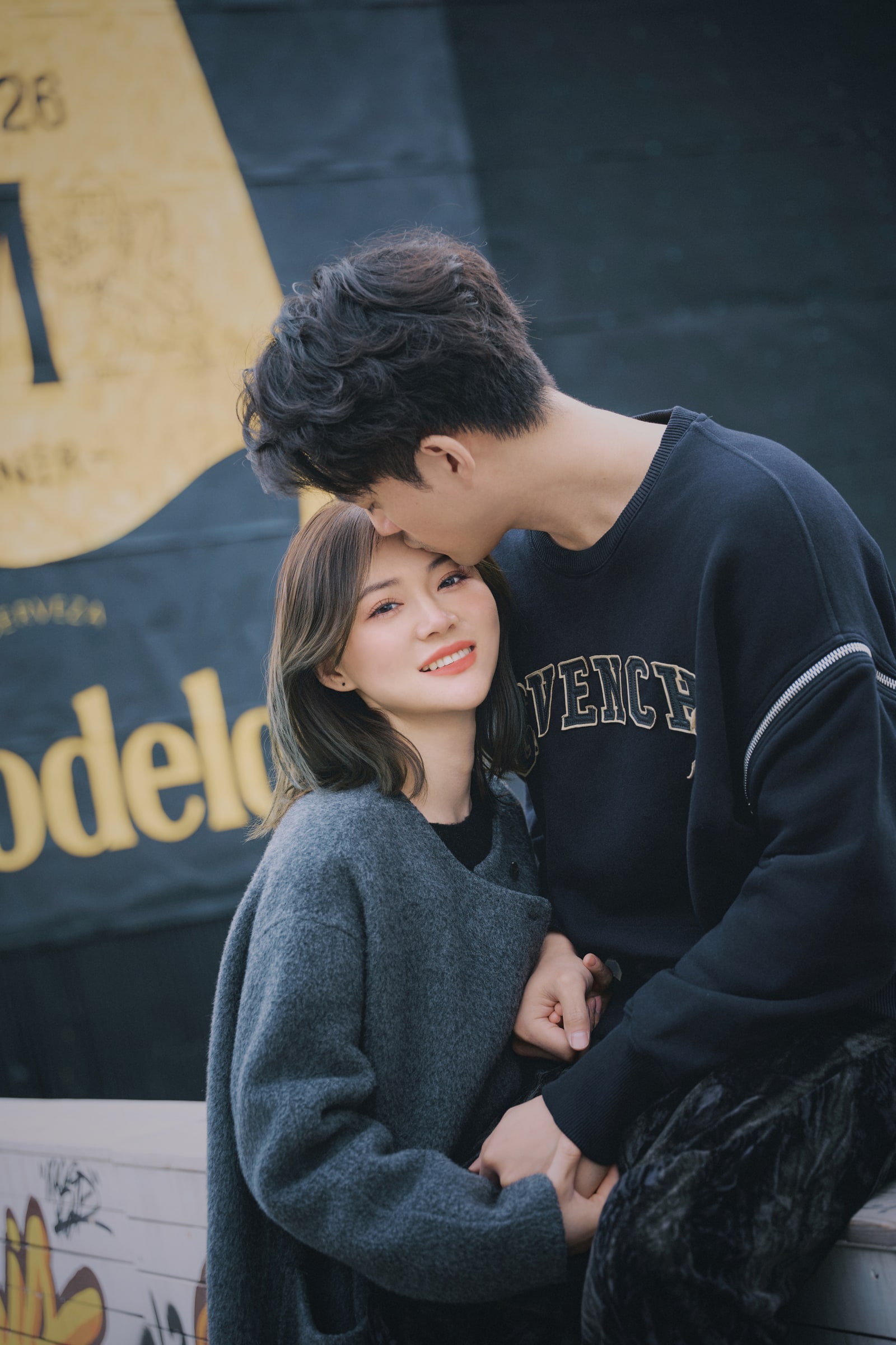 A couple holding hands as one kisses the other on the forehead beside a painted wall