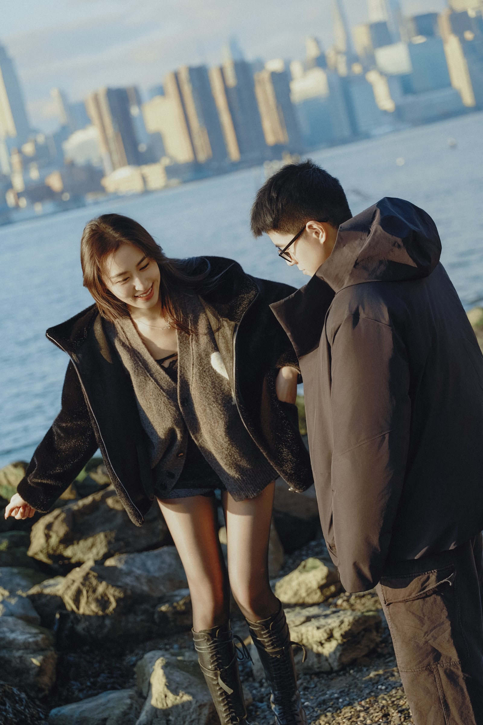 A couple walking over lakeside rocks with the city skyline across the water