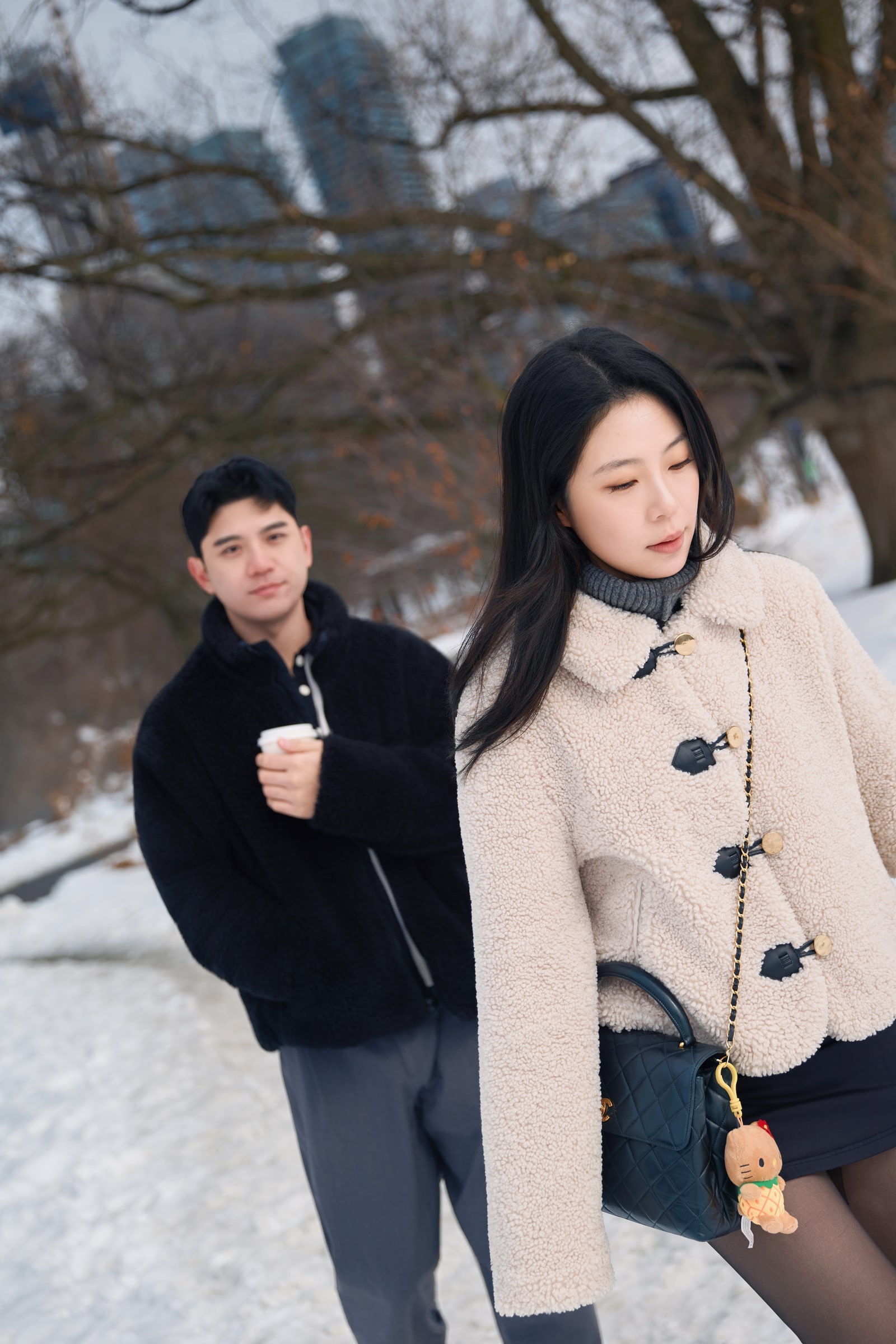 A couple in winter coats walking along a snowy path beneath bare trees