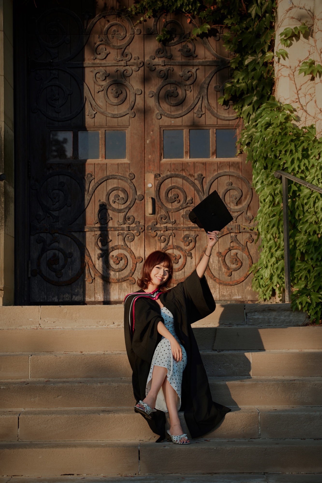 A graduate seated on sunlit stone steps, raising her cap beside an ivy-covered wooden door