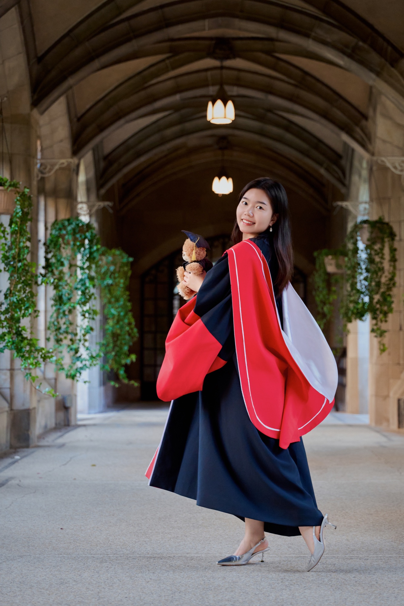 A graduate in red and navy doctoral robes holding a teddy bear beneath stone arches