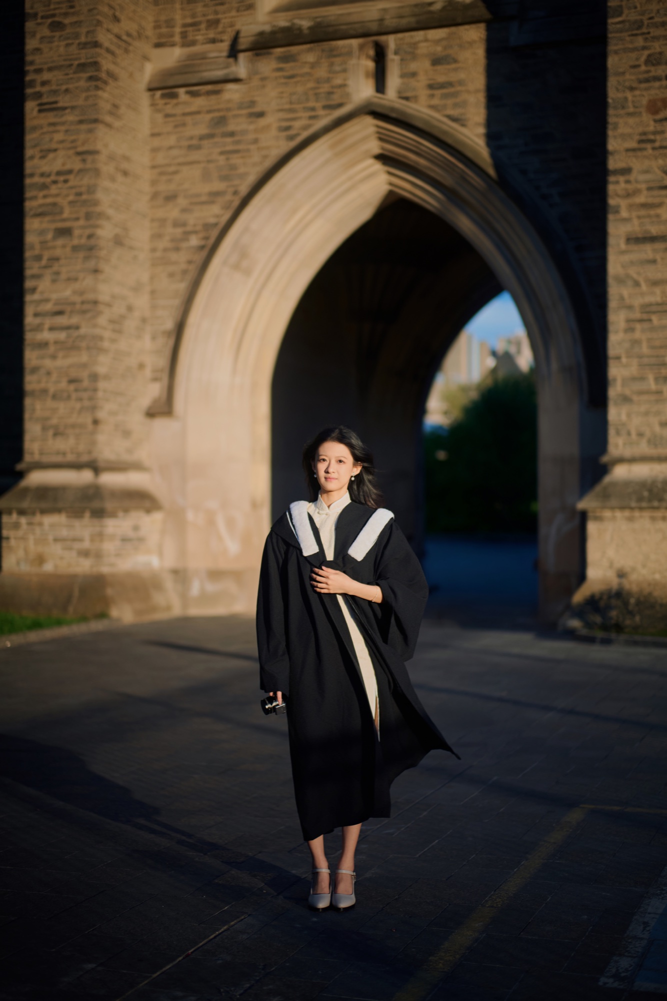 A graduate standing in evening sunlight in front of a tall stone arch