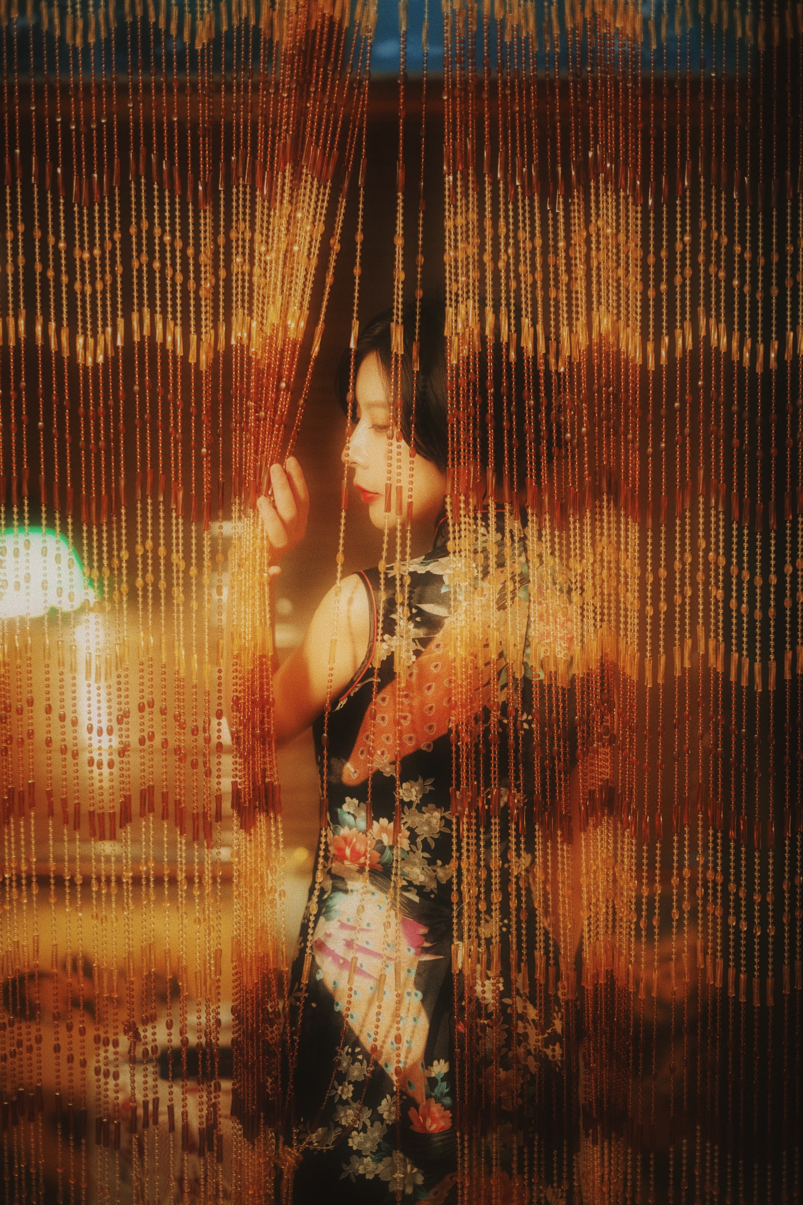 A portrait in a floral dress behind beaded curtains, illuminated by warm amber light