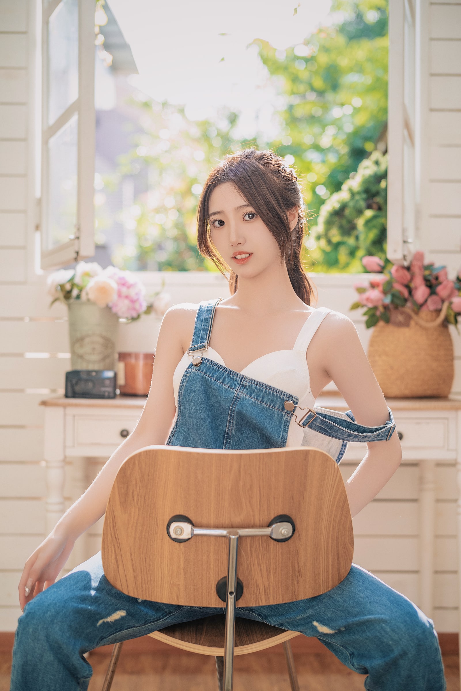 A portrait in denim overalls seated behind a wooden chair in a sunlit room
