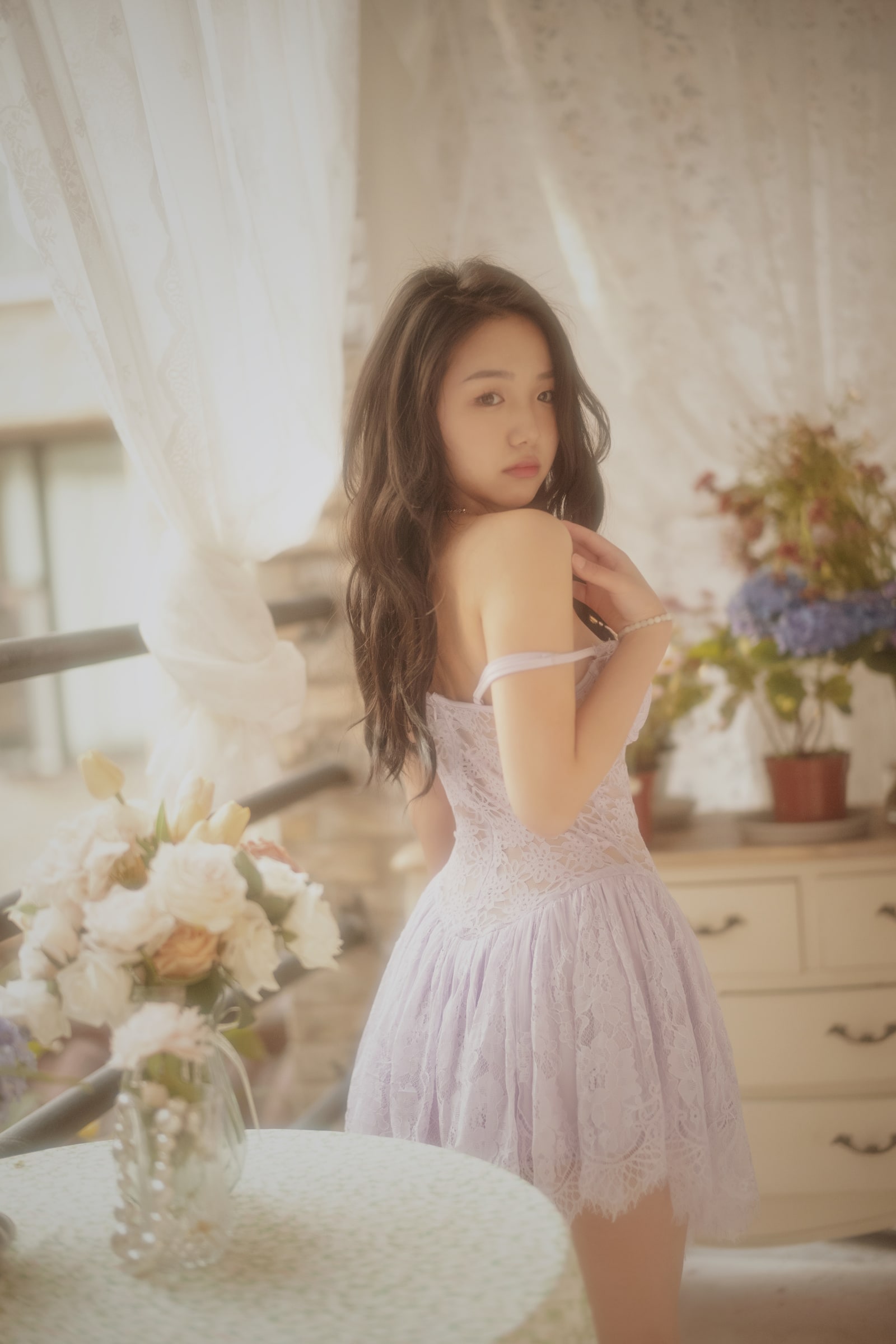 A portrait in a lilac lace dress, looking over one shoulder beside a sunlit window and flowers