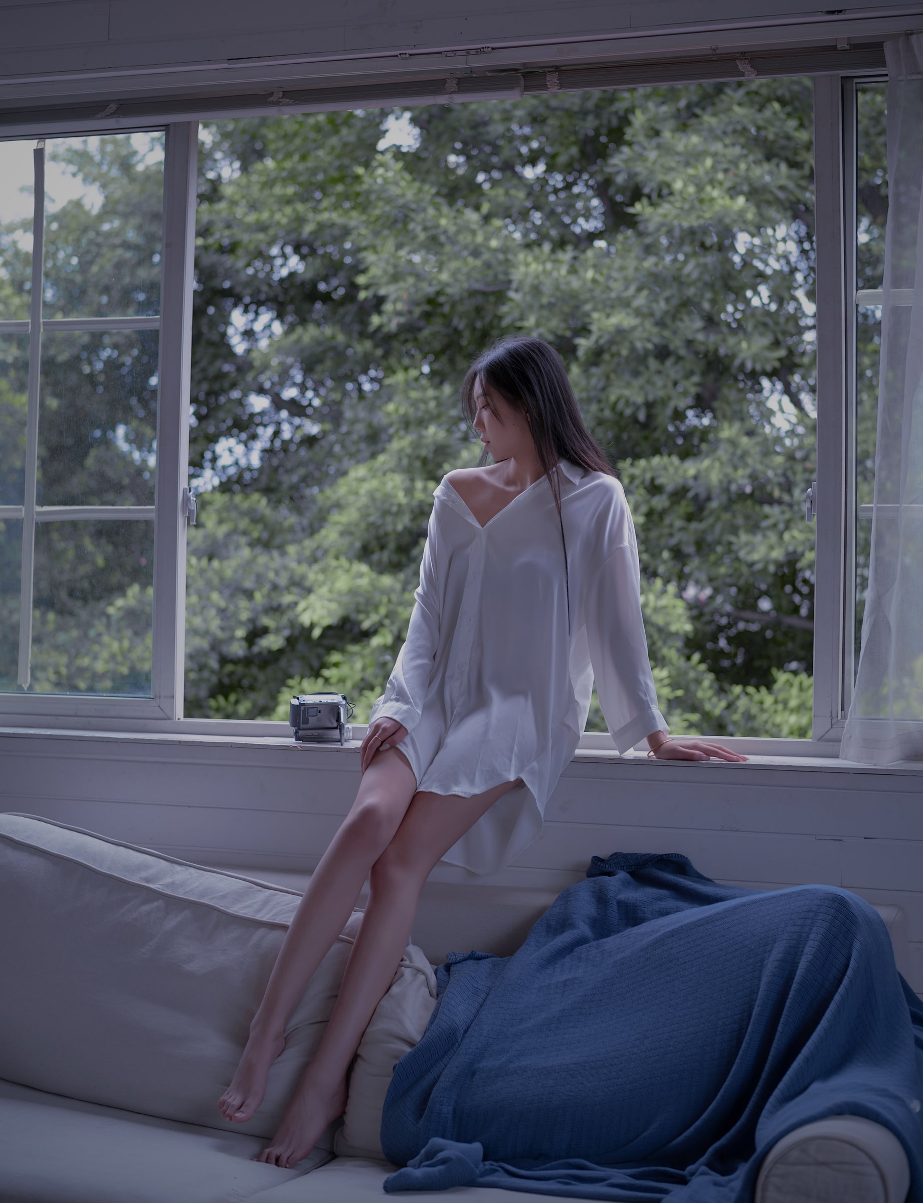 A portrait in a white shirt beside an open window overlooking green trees