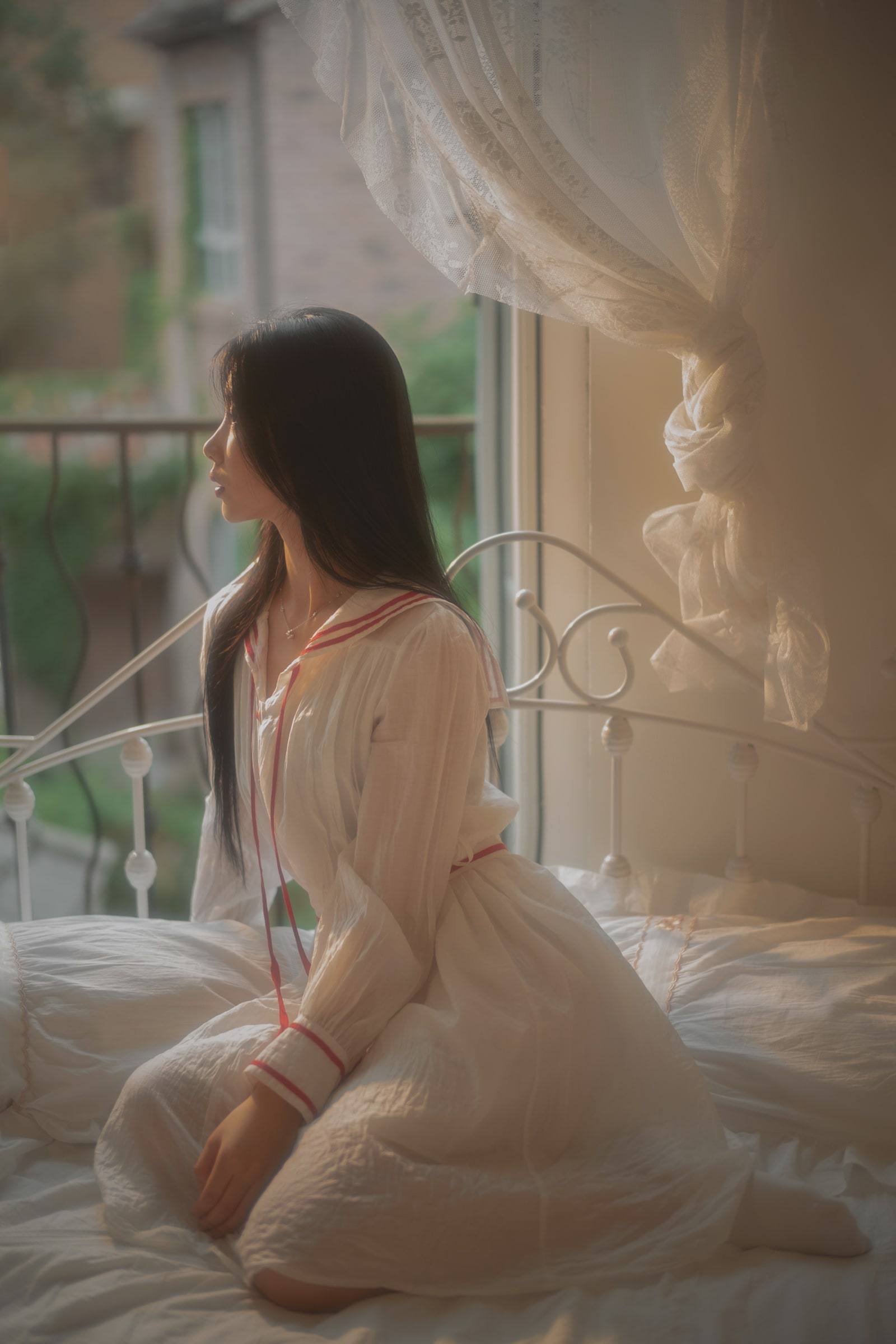 A side-profile portrait in a white dress, seated beside a softly lit window and lace curtains