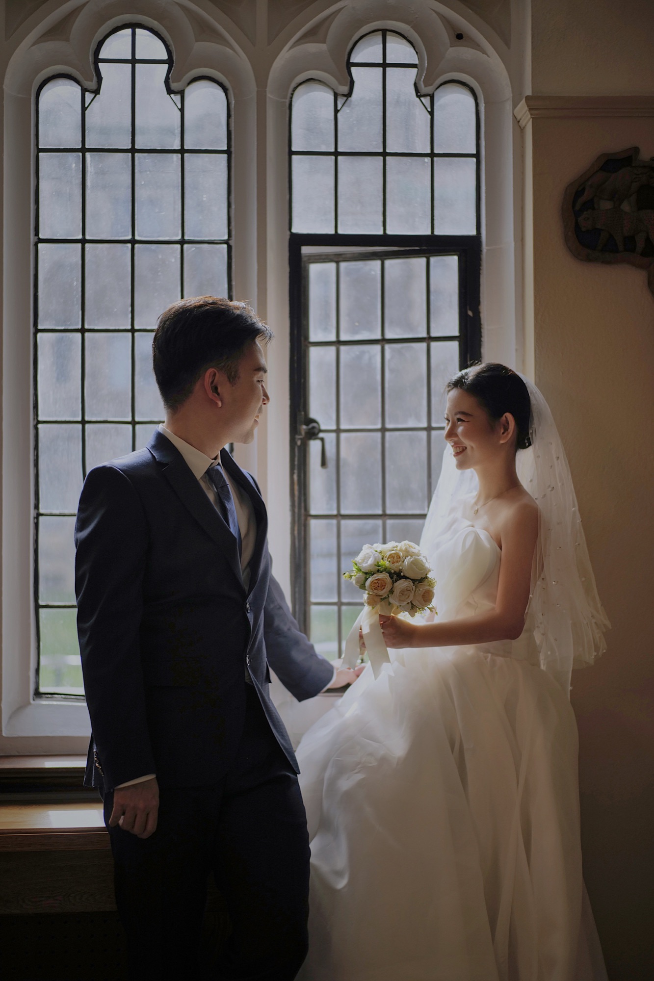 A wedding couple smiling beside tall windows