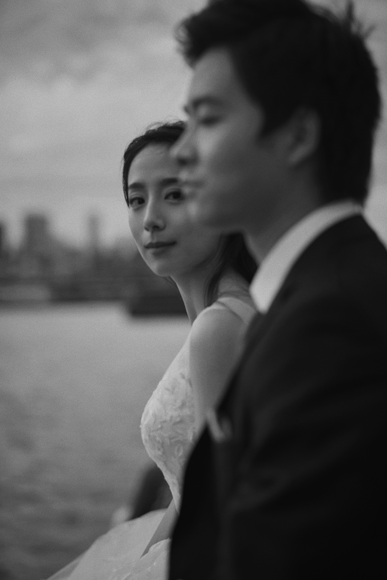 A black and white wedding portrait by the water, with the bride behind the softly blurred groom