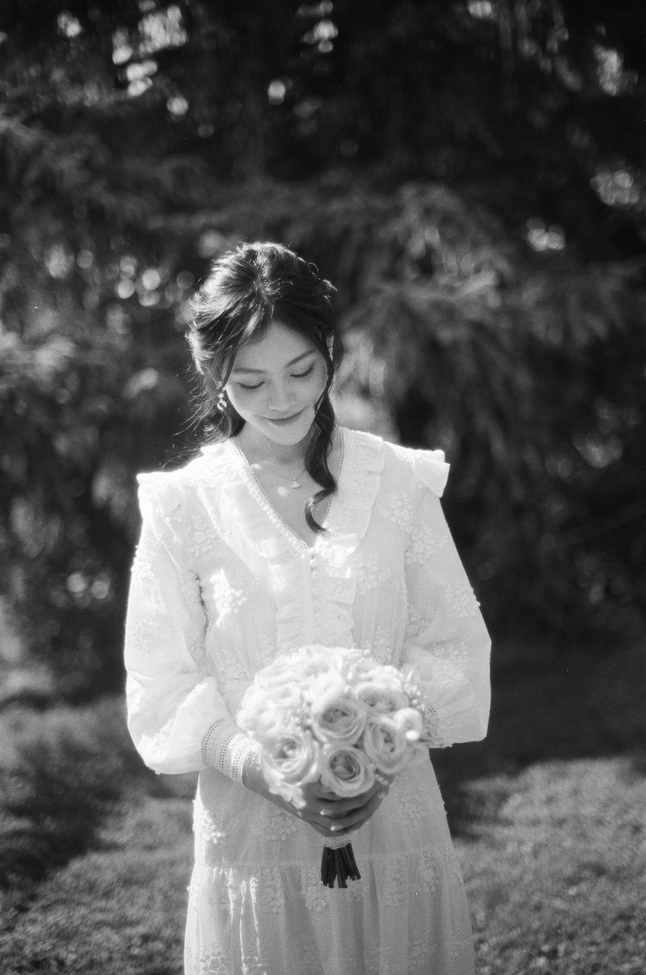 A black and white portrait of a bride looking down at her bouquet in a sunlit garden