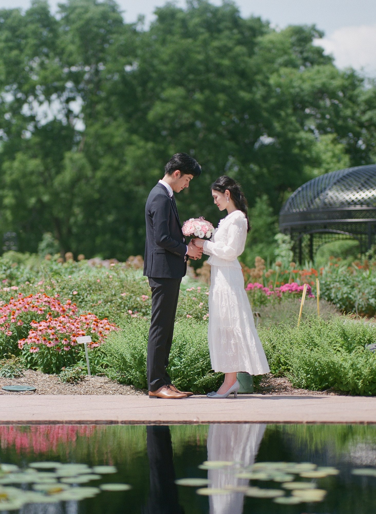 A wedding couple holding a pink bouquet in a flower garden, reflected in a pond