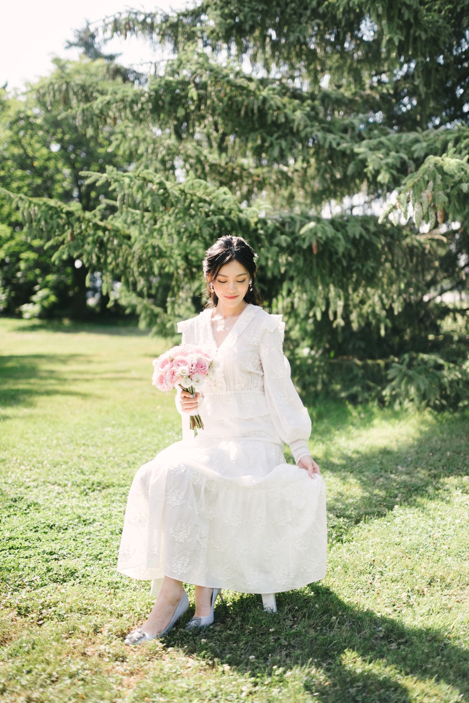 A seated bride holding a pink bouquet in a sunlit garden