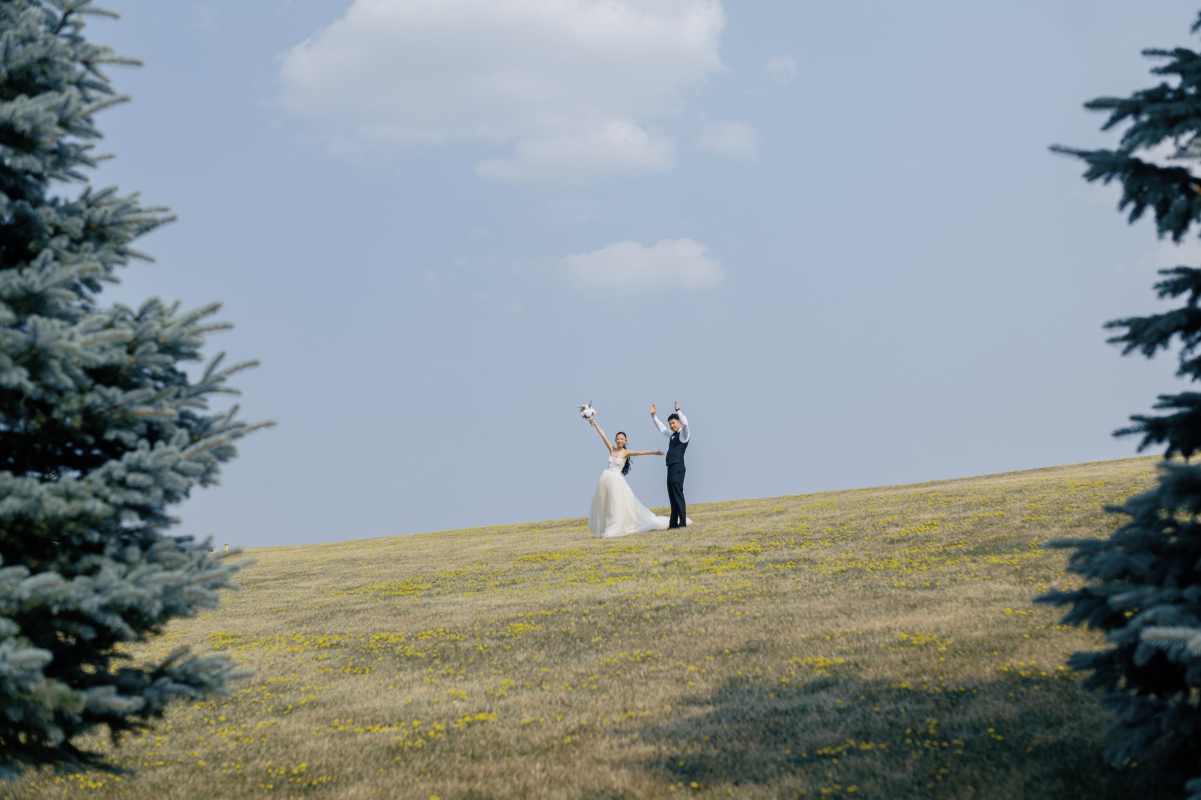 A wedding couple raising their arms on a flower-covered hillside beneath a wide blue sky