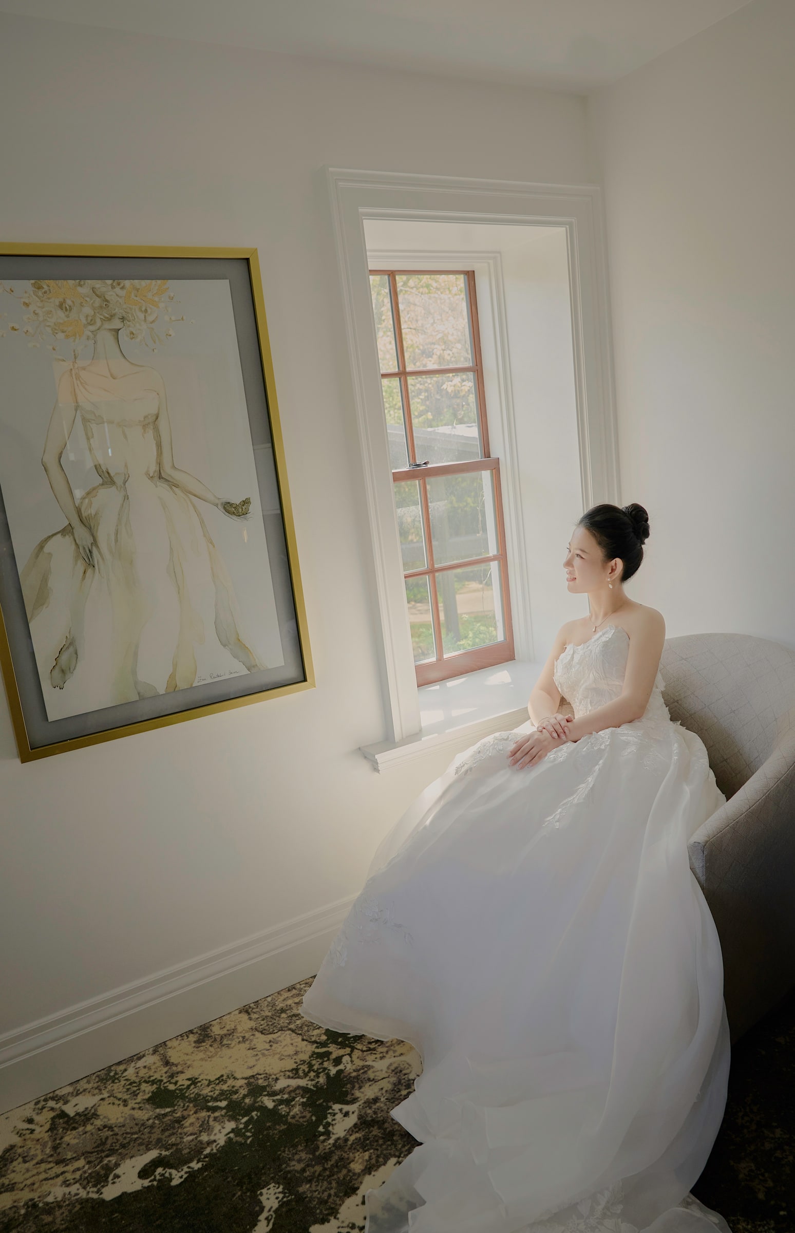 A bride seated beside a sunlit window and a framed bridal illustration
